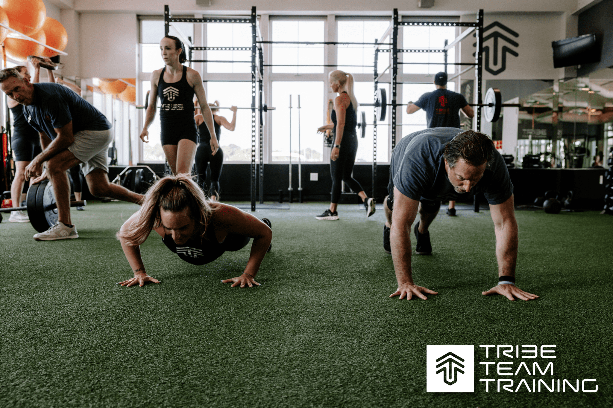 TRIBE Team Training group class at Inlet Fitness — participants and coaches performing push-ups and strength drills on turf in a coach-led small-group workout.